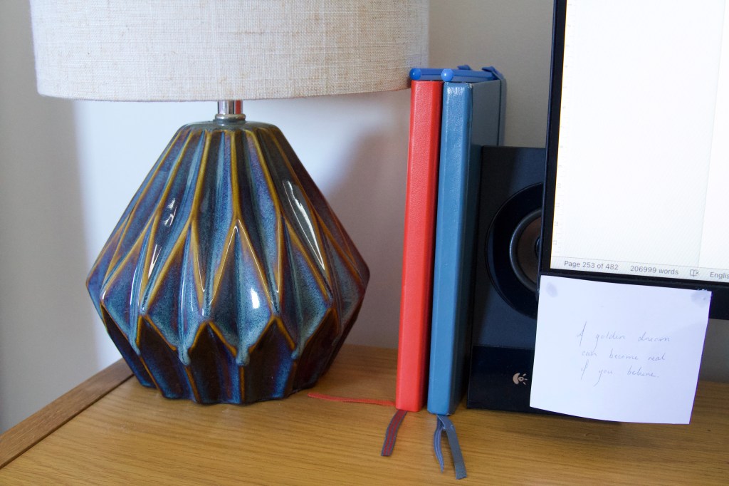 An orange and a blue notebook on a desk between a lamp and a speaker. A computer monitor is shown in the right corner.
